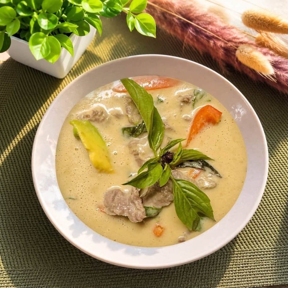 Bowl of creamy Thai green curry with meat, vegetables, and fresh basil garnish on a woven placemat.